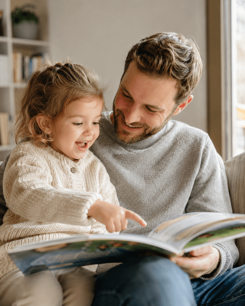 Elternteil und Kind schauen gemeinsam ein Bilderbuch an und sprechen miteinander, liebevolle Alltagsszene (KI-generiert)