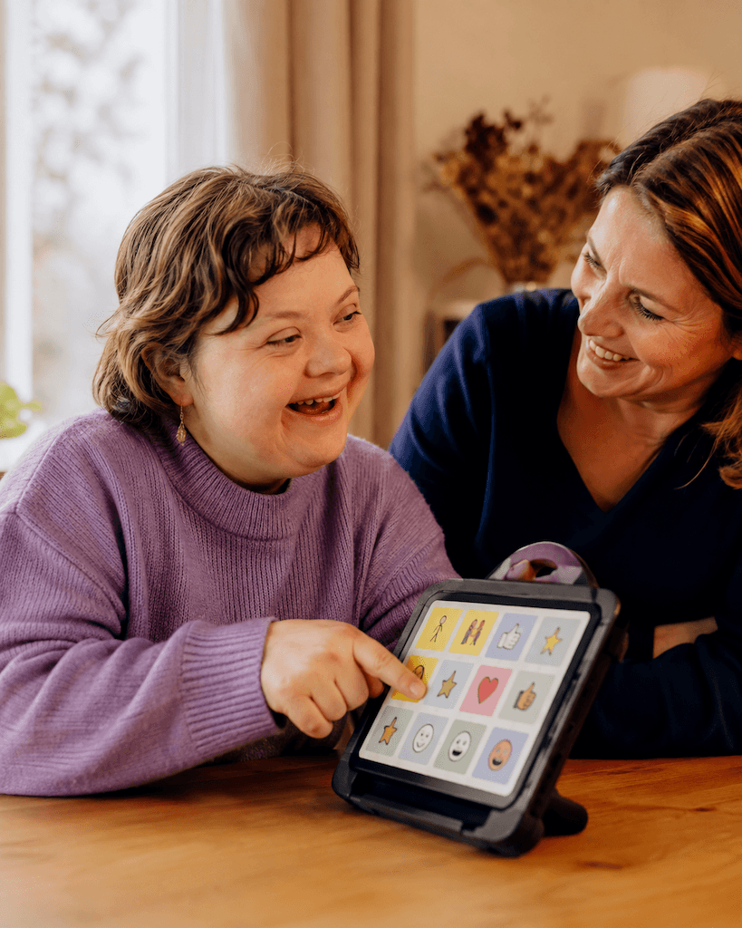 Mensch kommuniziert freudig mit unterstützter Kommunikation, etwa einer Bildtafel oder einem Tablet-Talker, mit einer zugewandten Person (KI-generiert)