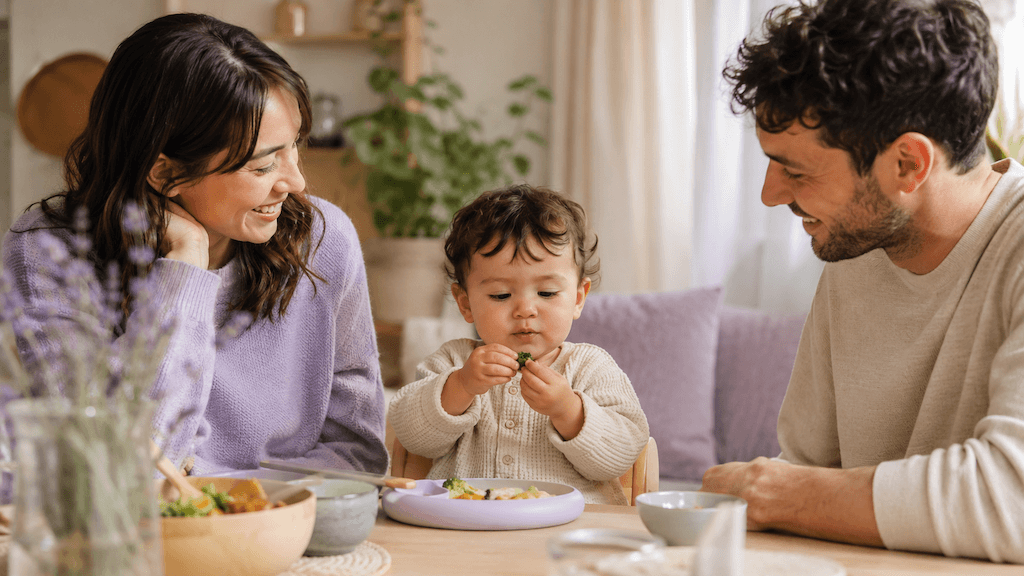 Kleinkind erkundet beim entspannten Familienessen neugierig ein Stück Essen, die Eltern bleiben gelassen und ohne Druck (KI-generiert)